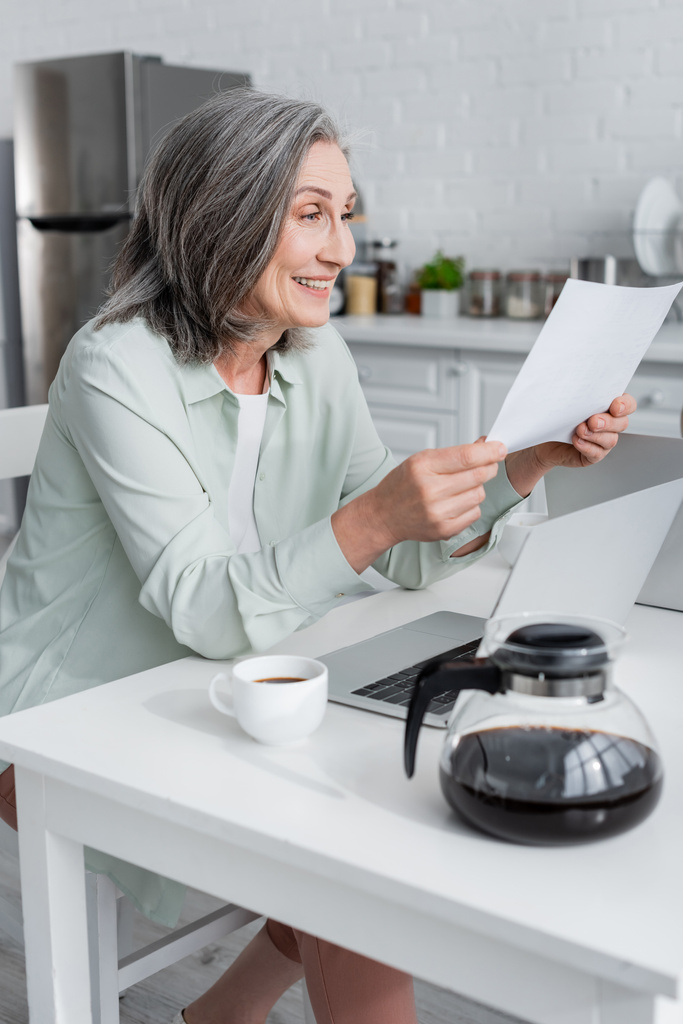 Wesoła kobieta trzyma papier w pobliżu kawy i laptopa w domu  - Zdjęcie, obraz