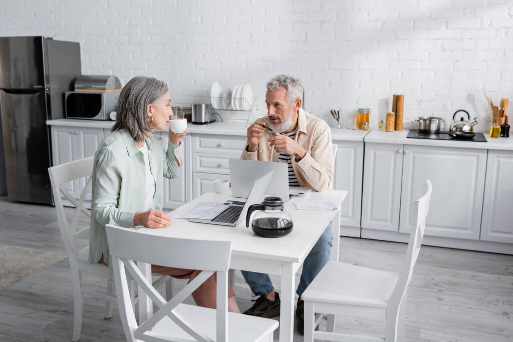Мужчина средних лет смотрит на жену с чашкой кофе возле счетов и ноутбуков на кухне  - Фото, изображение