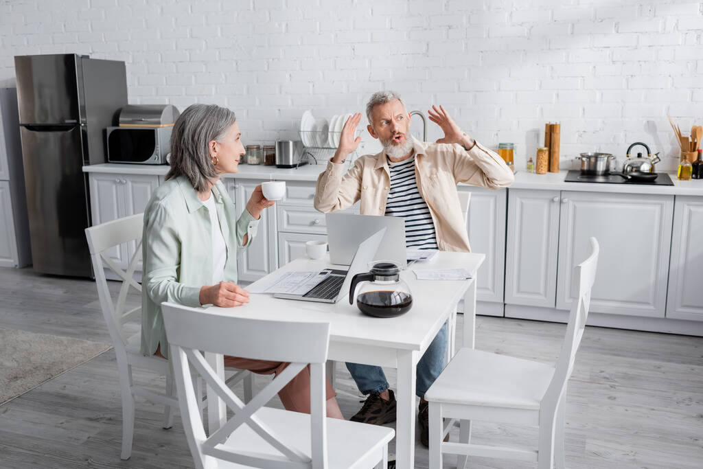 Зрелый мужчина разговаривает с женой с кофе возле газет и ноутбуков на кухне  - Фото, изображение