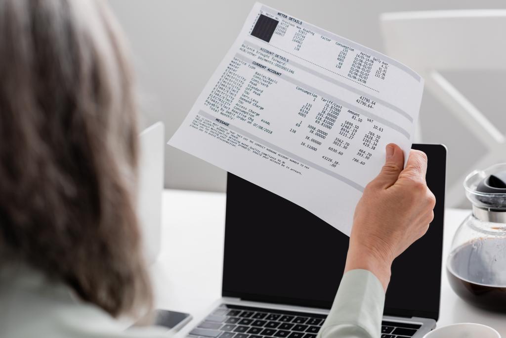 Papier avec des factures en main de femme mature près des ordinateurs portables et du café à la maison  - Photo, image