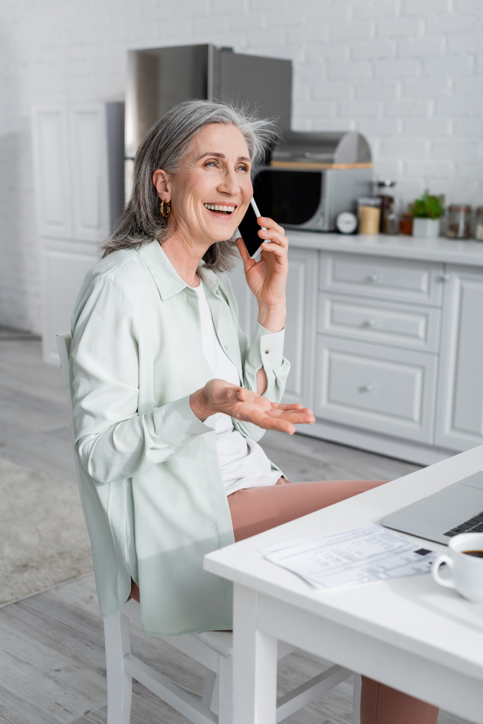 Mujer madura sonriente hablando en el teléfono inteligente cerca de la computadora portátil, café borroso y facturas en la cocina  - Foto, imagen