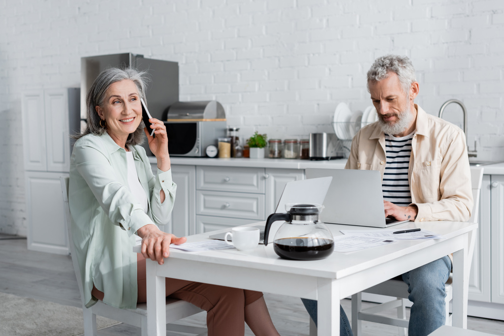 Kypsä nainen puhuu älypuhelimella lähellä miestä käyttäen kannettavaa tietokonetta ja laskuja pöydällä keittiössä  - Valokuva, kuva