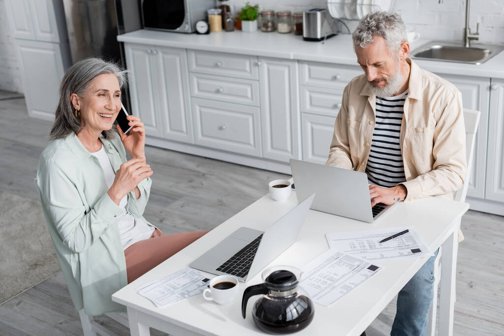 Nagy látószögű kilátás férfi használ laptop közelében kávé, számlák és feleség beszél okostelefon a konyhában  - Fotó, kép