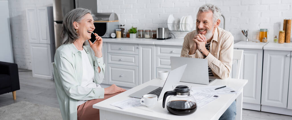 Mosolygós férfi nézi vidám feleség beszél a mobiltelefon közelében laptop, kávé és számlák a konyhában, banner  - Fotó, kép