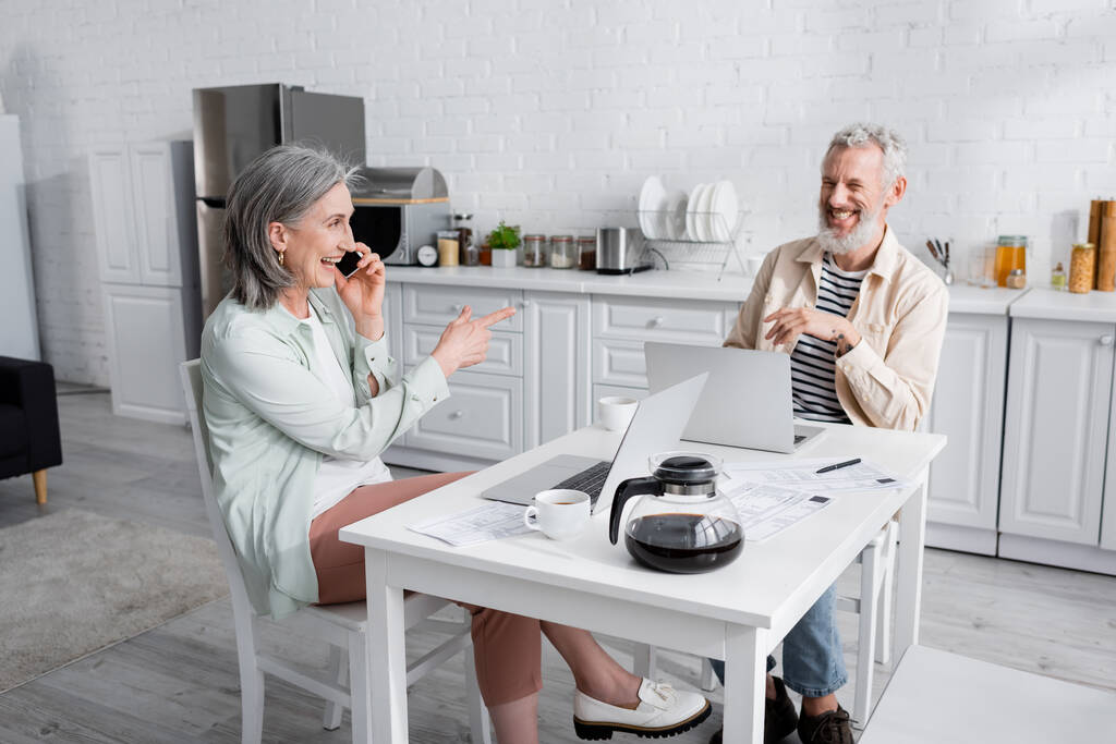 Mujer madura sonriente tomando el teléfono celular y señalando al marido cerca de computadoras portátiles y facturas en la cocina  - Foto, imagen