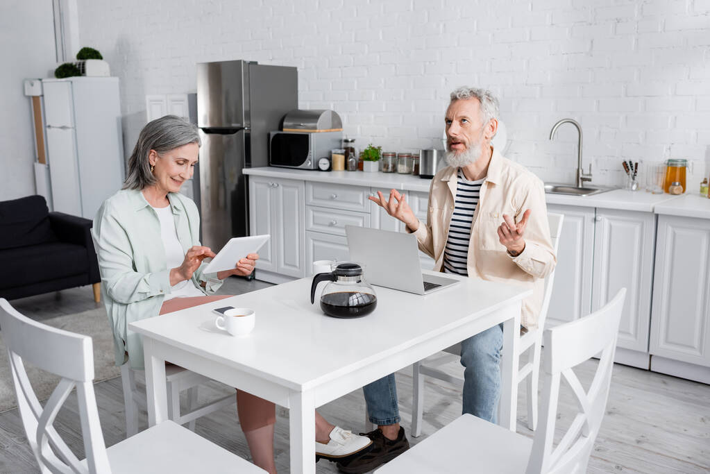 Pensive starší muž sedí v blízkosti notebooku, kávy a manželky s digitálním tabletem v kuchyni  - Fotografie, Obrázek