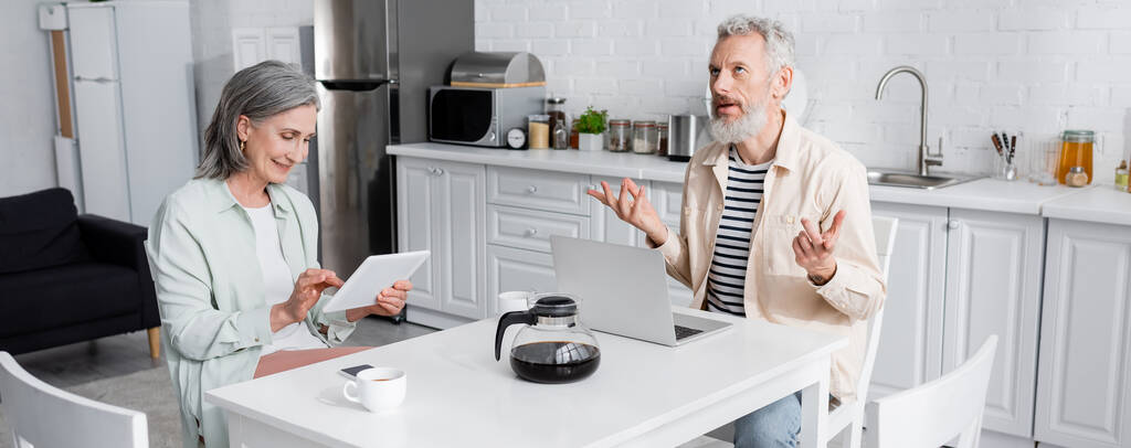 Sorrindo mulher usando tablet digital perto de marido maduro pensativo e café na cozinha, banner  - Foto, Imagem