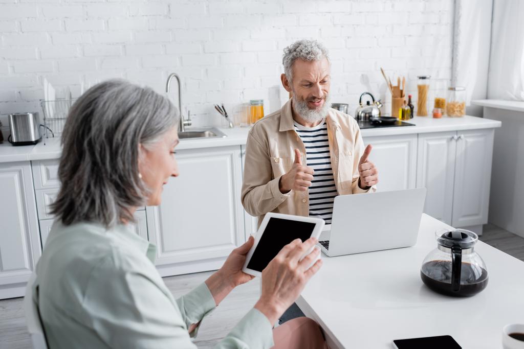 Зрелый мужчина показывает большие пальцы вверх во время видеозвонка на ноутбуке рядом с кофе и жена с цифровой планшет дома  - Фото, изображение