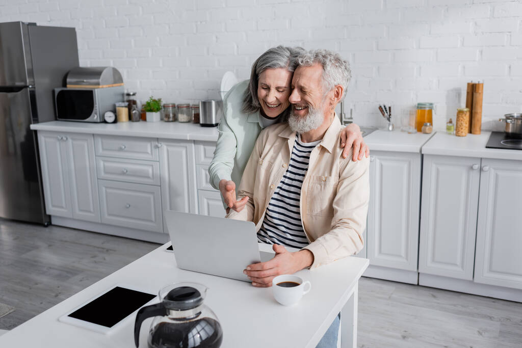 Χαρούμενη ώριμη γυναίκα αγκαλιάζει τον σύζυγο και δείχνει τον φορητό υπολογιστή κοντά στον καφέ και τις συσκευές στο σπίτι  - Φωτογραφία, εικόνα