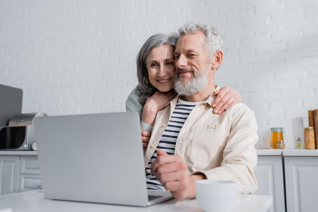 glimlachen volwassen vrouw knuffelen man in de buurt wazig laptop en beker in keuken  - Foto, afbeelding