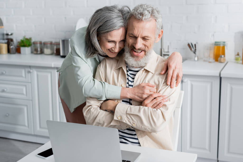volwassen vrouw knuffelen vrolijk echtgenoot op zoek naar laptop in de buurt smartphone in keuken  - Foto, afbeelding