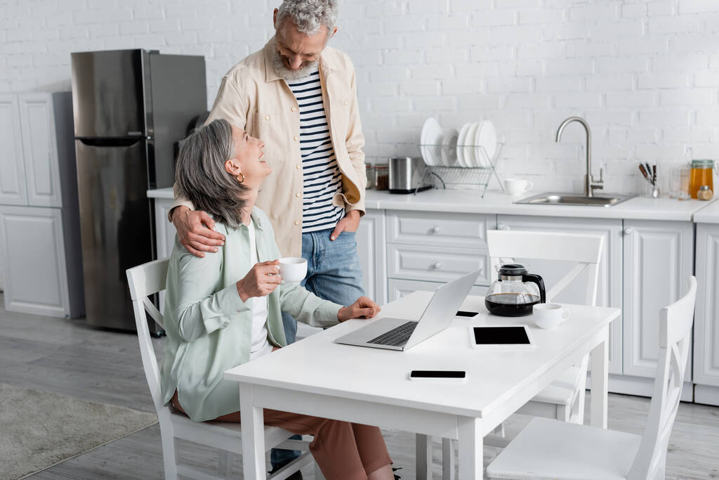 Homem alegre abraçando esposa com café perto de gadgets na cozinha  - Foto, Imagem