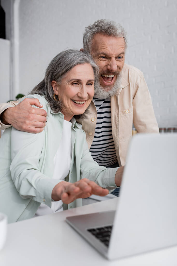 Homme excité étreignant femme mature près d'ordinateur portable flou à la maison  - Photo, image