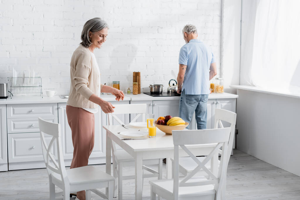 Reife Frau steht neben Orangensaft und Früchten auf Tisch in Küche  - Foto, Bild