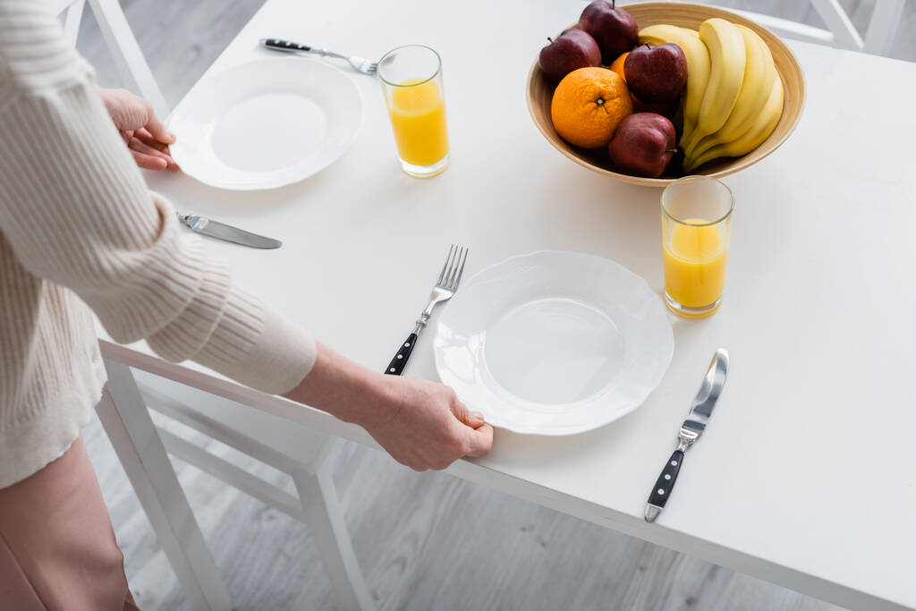 Ausgeschnittene Ansicht einer Frau, die Teller in der Nähe von Orangensaft und Früchten in der Küche auf den Tisch stellt  - Foto, Bild