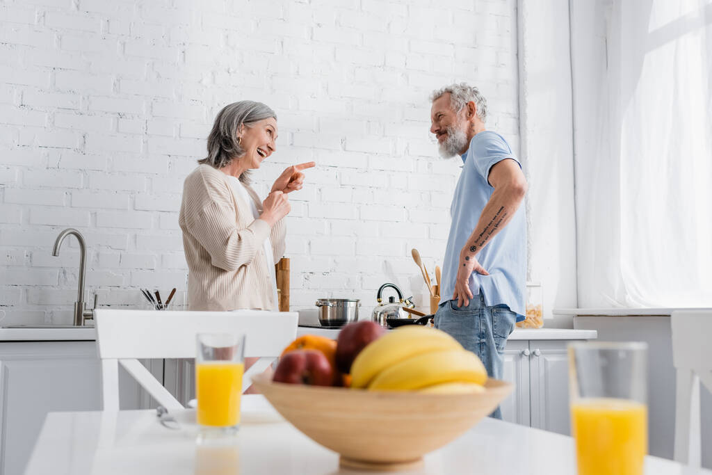 Uśmiechnięta kobieta wskazująca męża w pobliżu kuchenki i rozmazane owoce w kuchni  - Zdjęcie, obraz