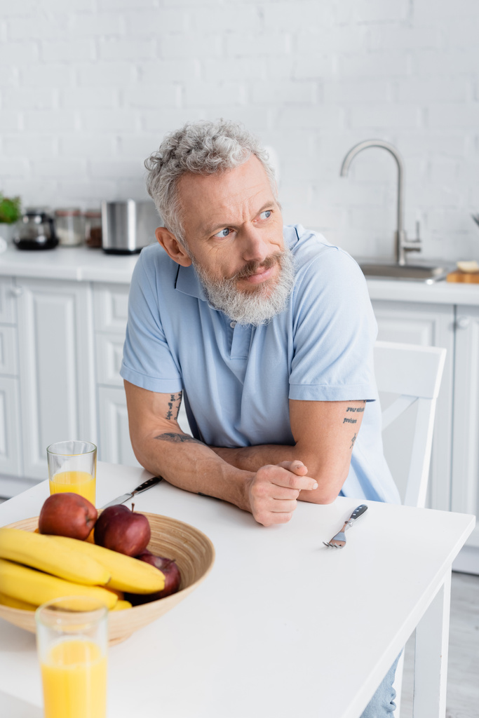 Uomo di mezza età seduto vicino a frutta e succo d'arancia in cucina  - Foto, immagini