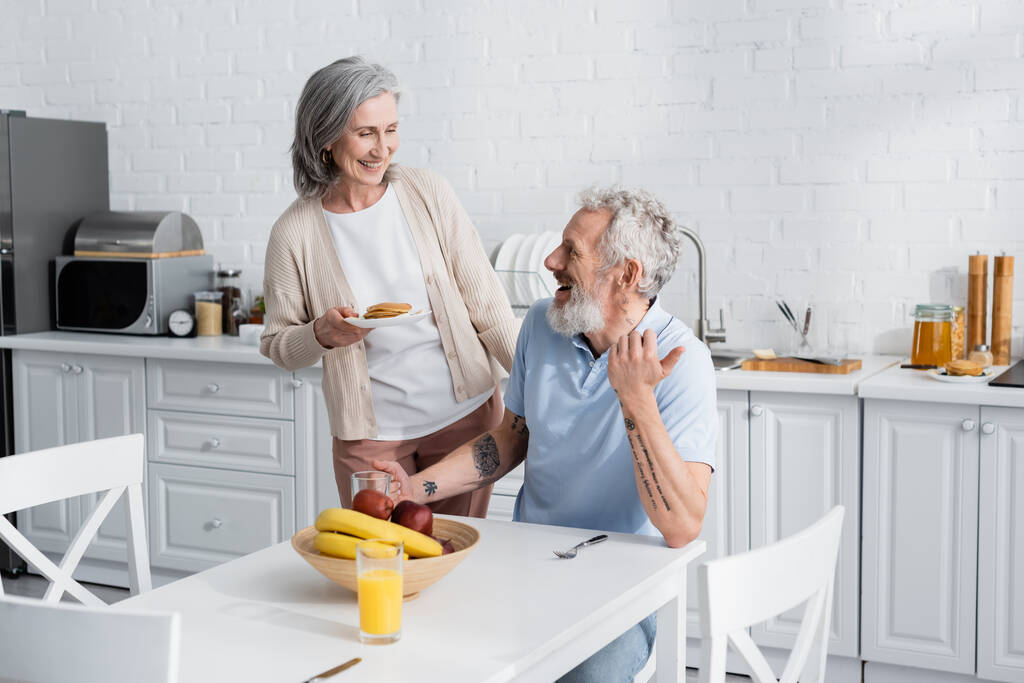 Χαμογελαστή γυναίκα κρατώντας τηγανίτες κοντά χαρούμενα σύζυγος και φρούτα στην κουζίνα  - Φωτογραφία, εικόνα