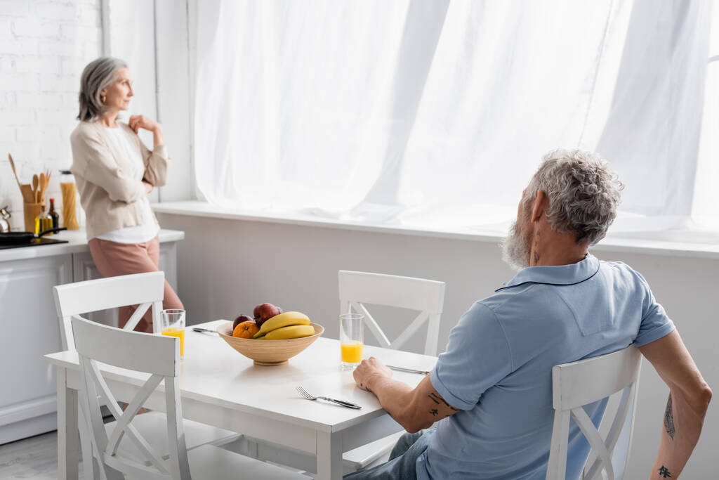 Mature homme assis près de jus d'orange et femme floue dans la cuisine  - Photo, image