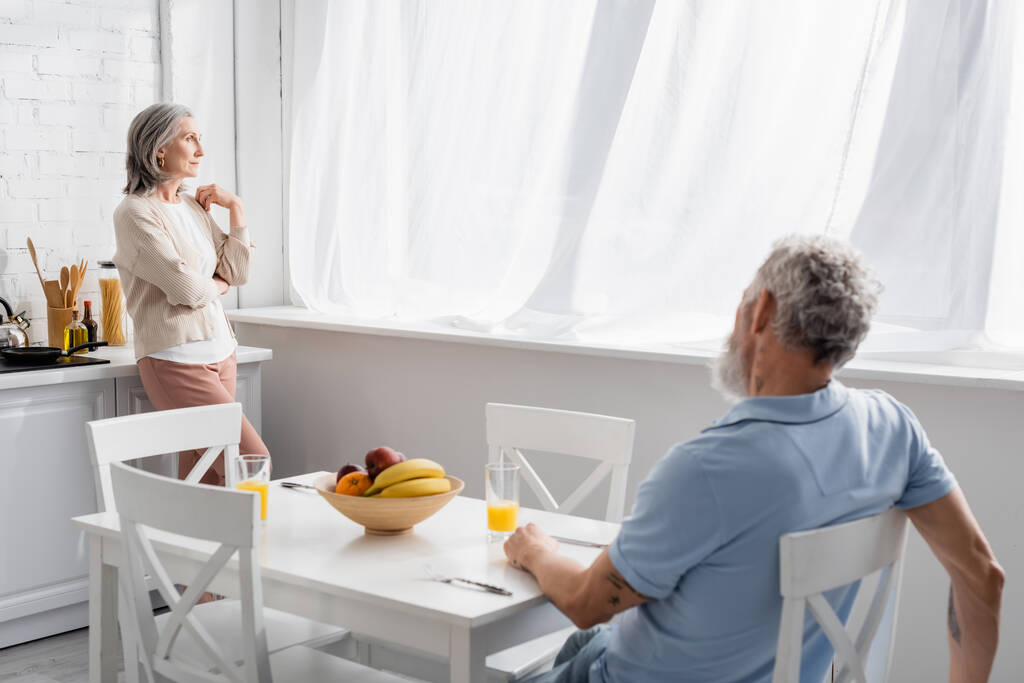 Μεσήλικη γυναίκα που κοιτάζει το παράθυρο κοντά στο σύζυγο στην κουζίνα  - Φωτογραφία, εικόνα