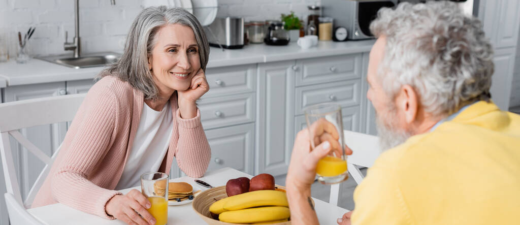 Mujer de mediana edad sosteniendo un vaso de jugo de naranja cerca del desayuno y esposo borroso, pancarta  - Foto, Imagen
