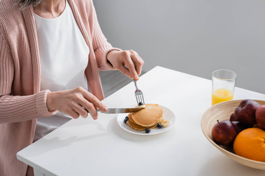 Обрізаний вид жінки, що ріже млинці біля свіжих фруктів і апельсиновий сік на кухні
  - Фото, зображення