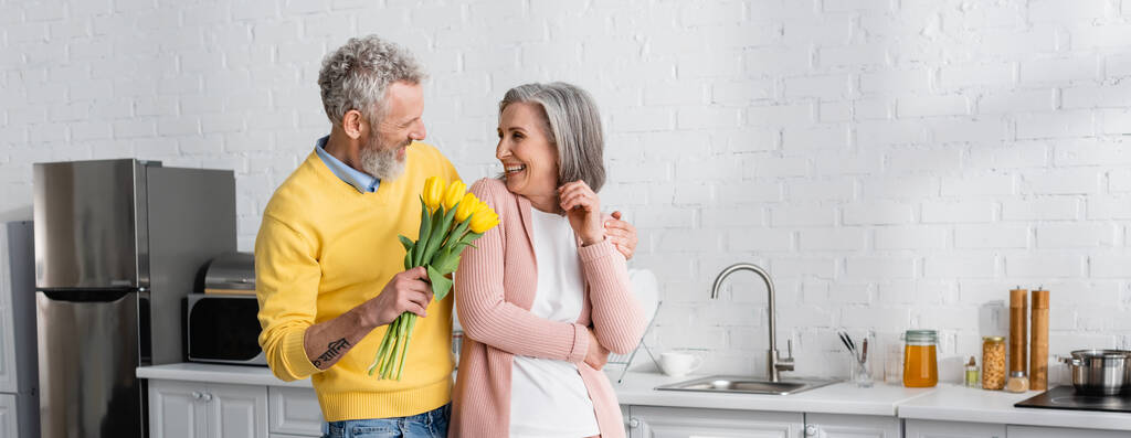 Lächelnder Mann umarmt Frau und hält gelbe Tulpen in der Küche, Transparent. Übersetzung: "om, shanti, peace" - Foto, Bild