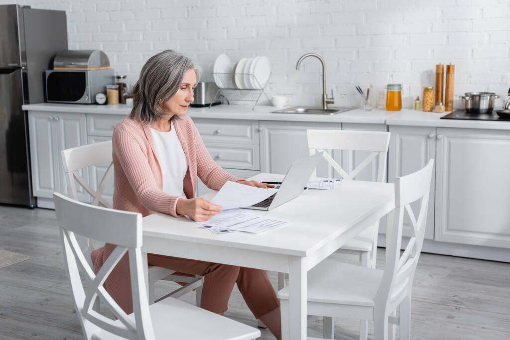 Mujer de mediana edad utilizando el ordenador portátil cerca de papeles con facturas en la cocina  - Foto, imagen