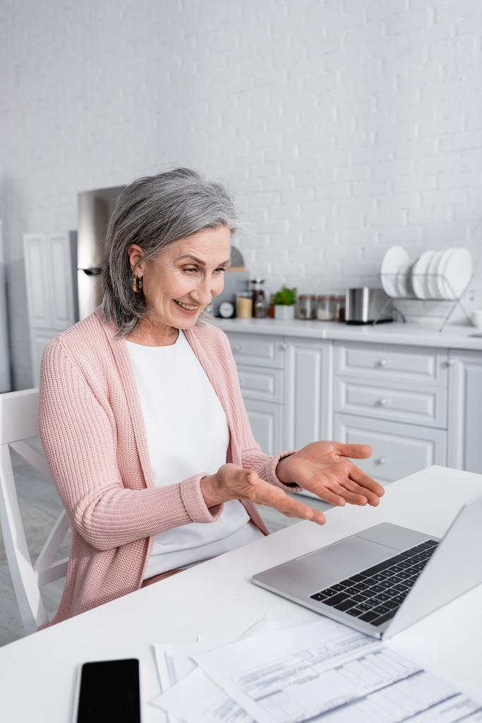 Mujer de mediana edad positiva apuntando a la computadora portátil durante la videollamada cerca de facturas en casa  - Foto, Imagen