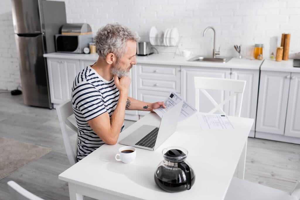 Вид збоку бородатого чоловіка, який дивиться на купюри біля ноутбука та кави на кухні
  - Фото, зображення