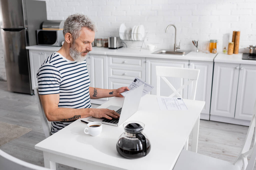 Evdeki kahvelerin yanında faturaları sayarken dizüstü bilgisayar kullanan bir adam. Çeviri: "om, shanti, peace" - Fotoğraf, Görsel