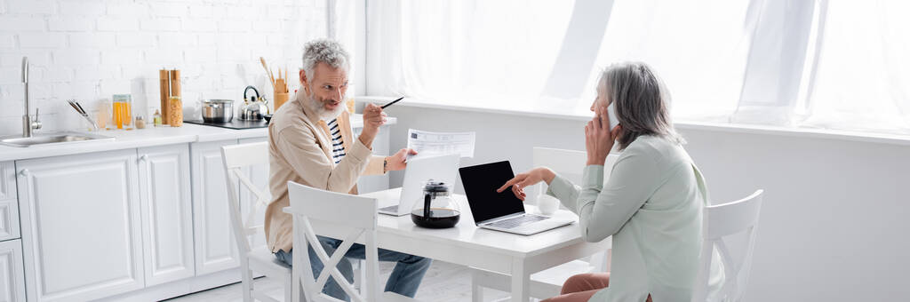 Férfi mutató feleség beszél okostelefon közelében számlák és laptopok a konyhában, banner  - Fotó, kép