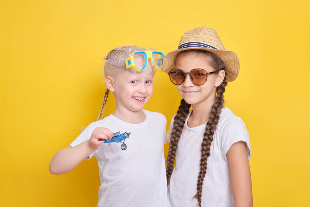dos chicas lindas esperando un viaje a la costa del mar, niña con una máscara para bucear y un avión de juguete, chica adolescente en un sombrero y gafas de sol, fondo amarillo - Foto, imagen