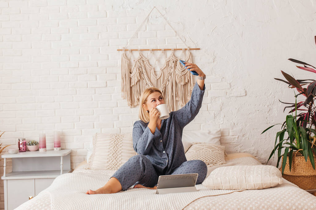 Kaunis nuori nainen pyjamassa, työskentelee kannettavan kanssa sängyssä, puhuu puhelimessa. Verkkokauppa - Valokuva, kuva