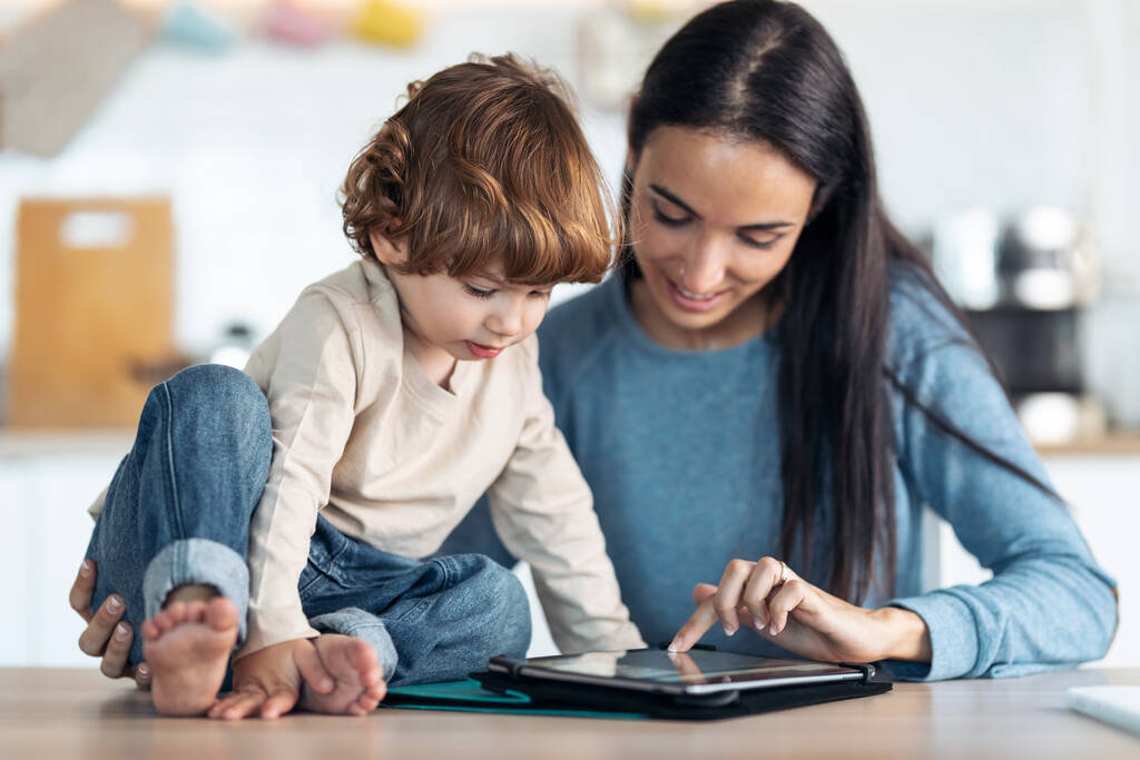 Aufnahme einer jungen lächelnden Mutter und ihres niedlichen Sohnes, die zu Hause in der Küche Cartoons auf einem digitalen Tablet ansehen. - Foto, Bild