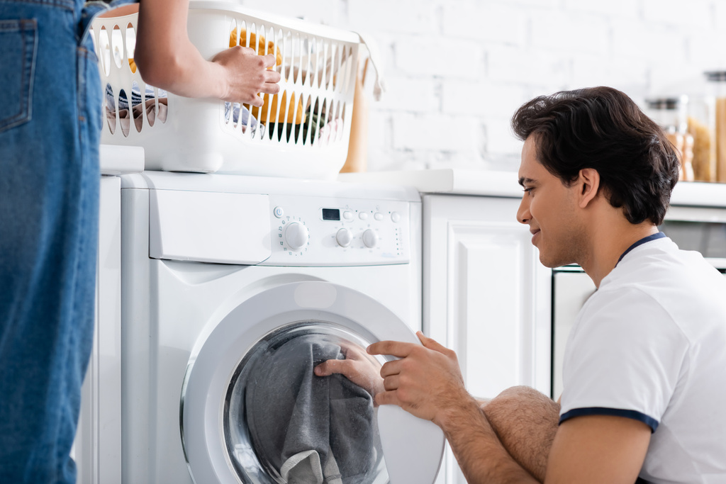 アフリカ系アメリカ人のガールフレンドの近くに洗濯機をロードする笑顔汚い洗濯のバスケット  - 写真・画像