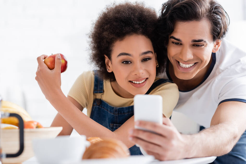 χαρούμενο διαφυλετικό ζευγάρι κοιτάζοντας smartphone κατά τη διάρκεια του πρωινού  - Φωτογραφία, εικόνα
