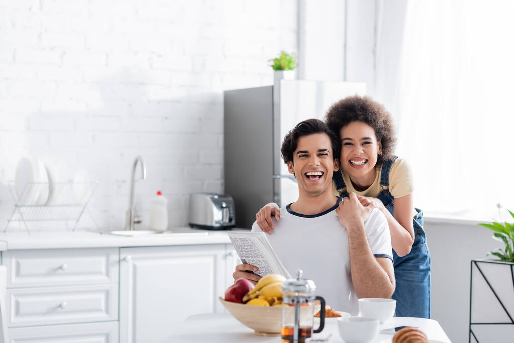 felice donna afroamericana che abbraccia fidanzato con giornale in cucina - Foto, immagini