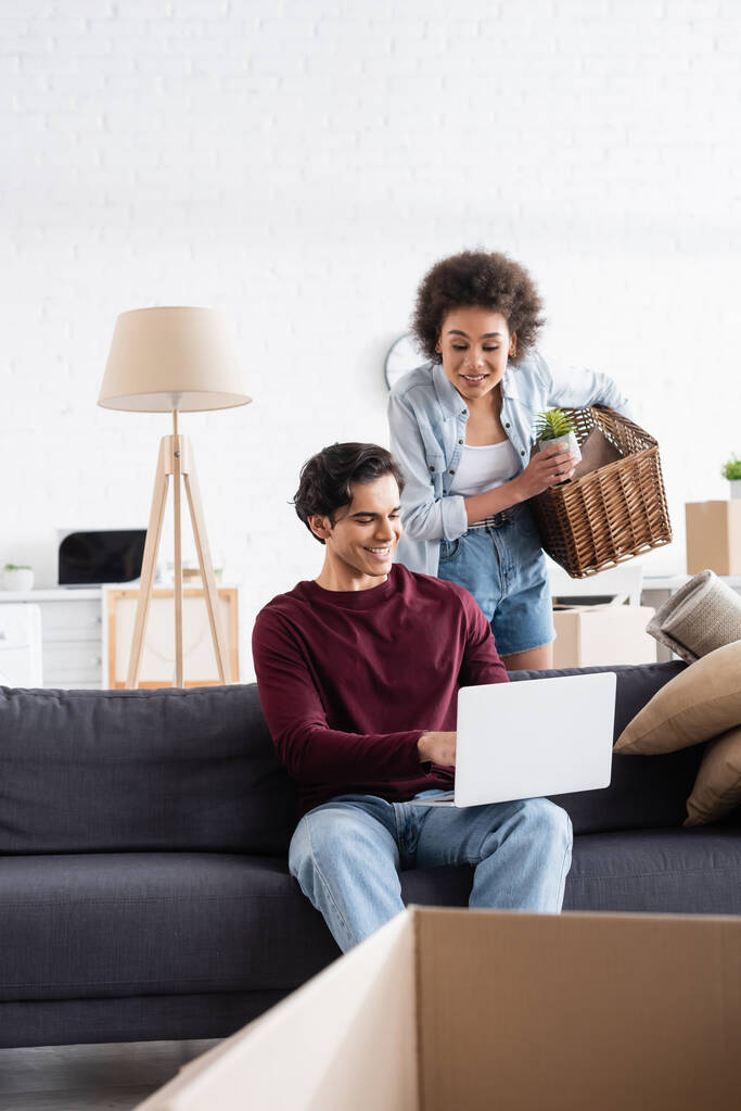 happy african american woman holding basket near boyfriend using laptop on couch - Photo, Image