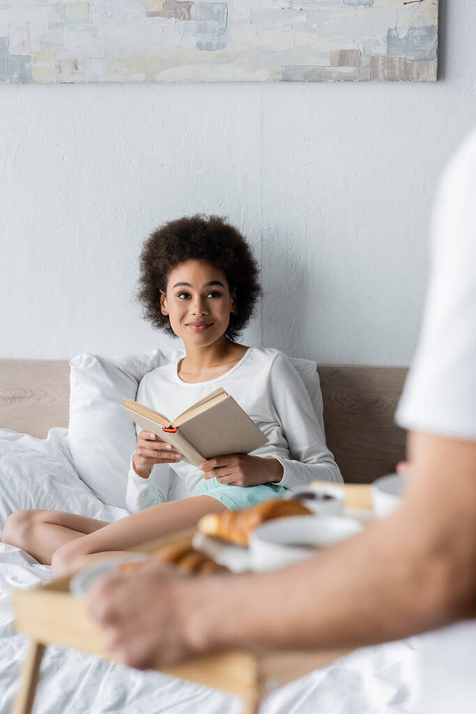zufrieden afrikanisch-amerikanische Frau sitzt auf dem Bett mit Buch in der Nähe verschwommenen Mann hält Frühstückstablett  - Foto, Bild