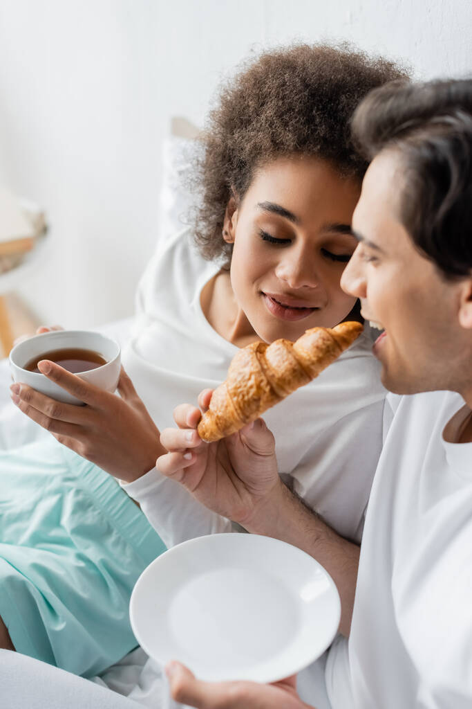 radosna międzyrasowa para jedząca śniadanie w łóżku  - Zdjęcie, obraz