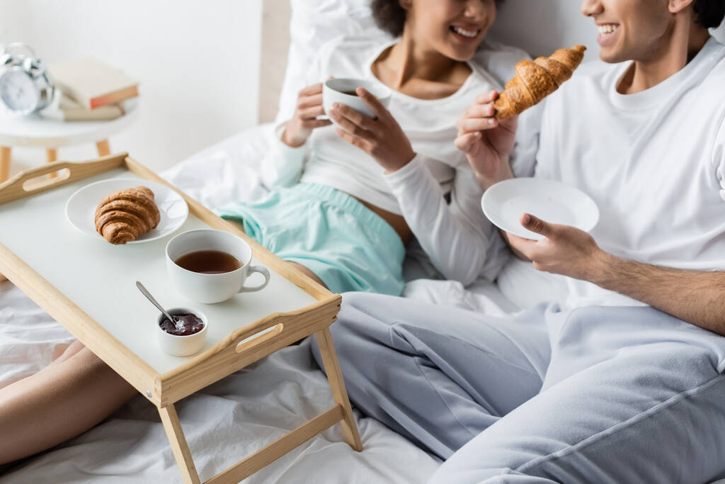 przycięty widok wesoły międzyrasowy para mając śniadanie w łóżku  - Zdjęcie, obraz