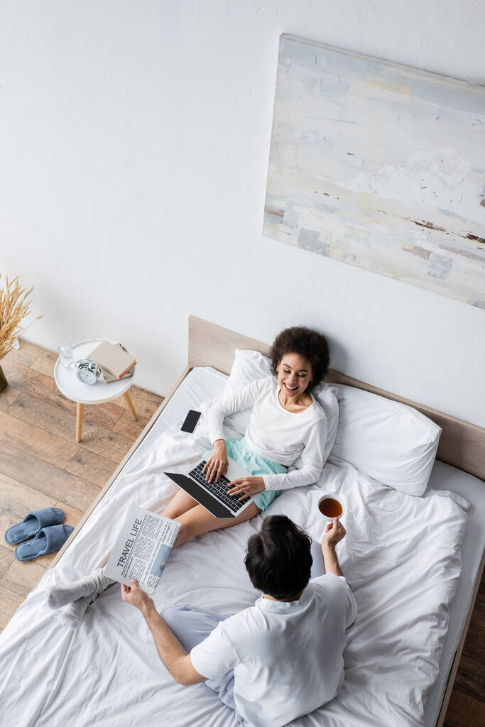 vue du haut de heureux afro-américain pigiste en utilisant un ordinateur portable près du petit ami tenant le journal de la vie de voyage dans le lit - Photo, image