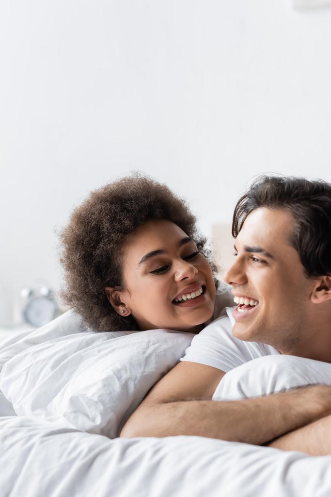 χαρούμενο διαφυλετικό ζευγάρι χαμογελά ενώ βρίσκεται στο κρεβάτι - Φωτογραφία, εικόνα