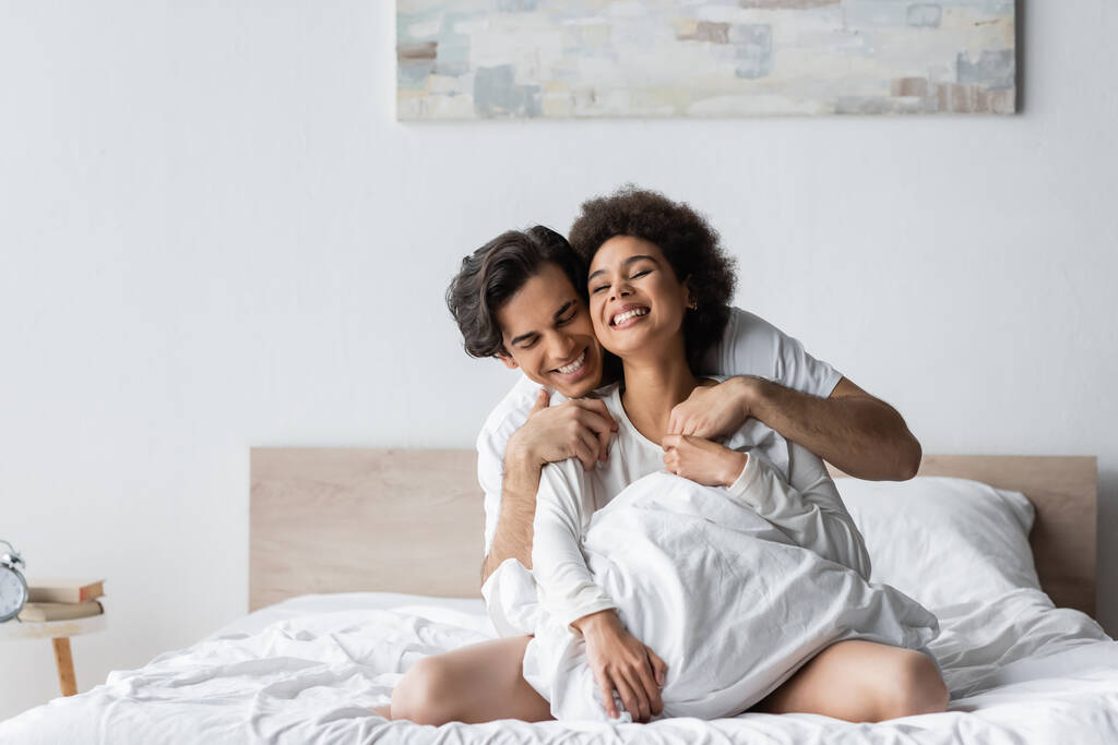 θετικό διαφυλετικό ζευγάρι χαμογελά ενώ αγκαλιάζει στο κρεβάτι - Φωτογραφία, εικόνα