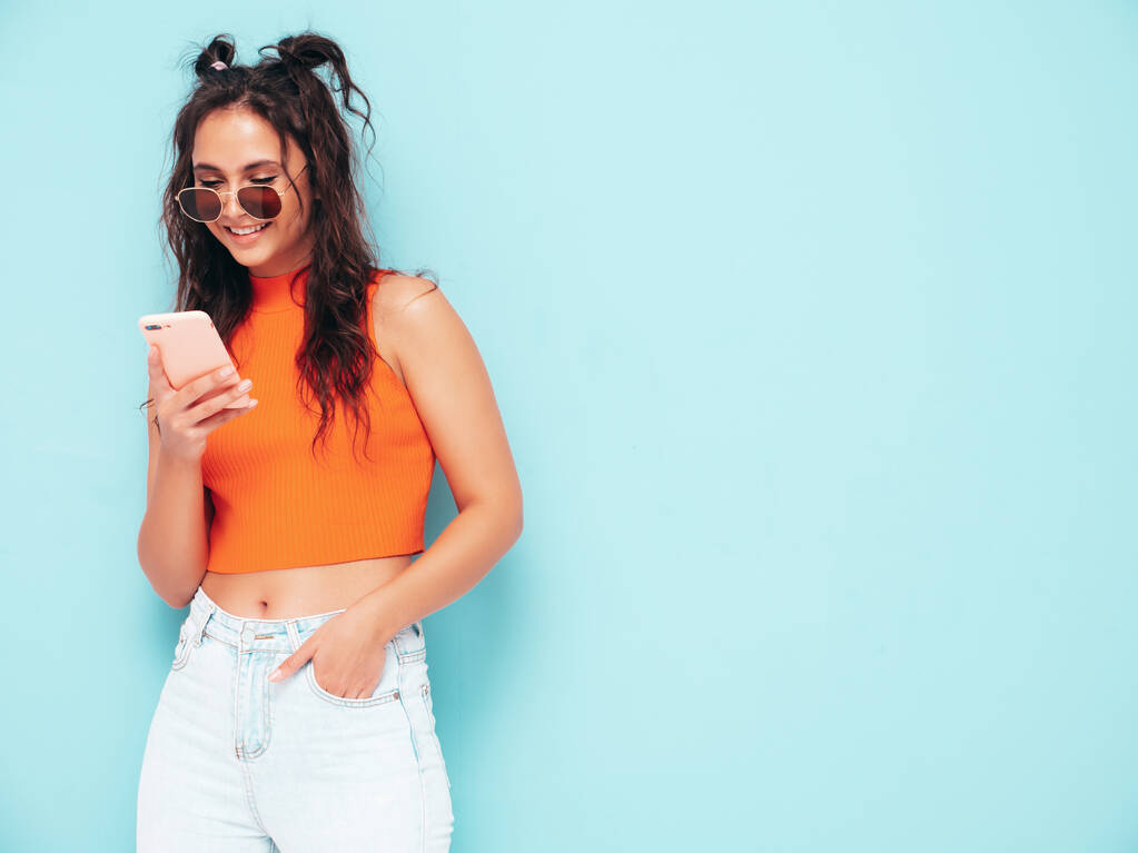 Молода красива усміхнена жінка в модному літньому оранжевому верхньому одязі та джинсовому одязі. Сексуальна безтурботна жінка позує біля синьої стіни в студії. Дивлячись на екран смартфона. Модель з використанням додатків для мобільних телефонів
 - Фото, зображення