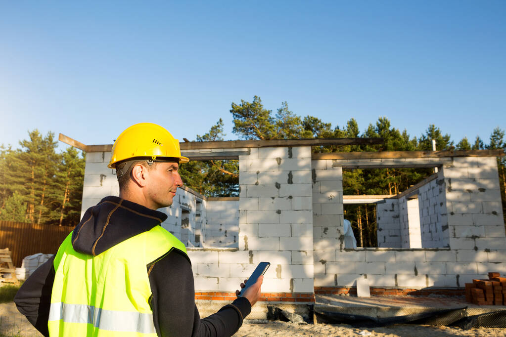 Trabalhador da construção fala em um smartphone em um chapéu amarelo e colete reflexivo contra o fundo da construção da casa as paredes e aberturas das janelas são feitas de blocos de concreto poroso. - Foto, Imagem