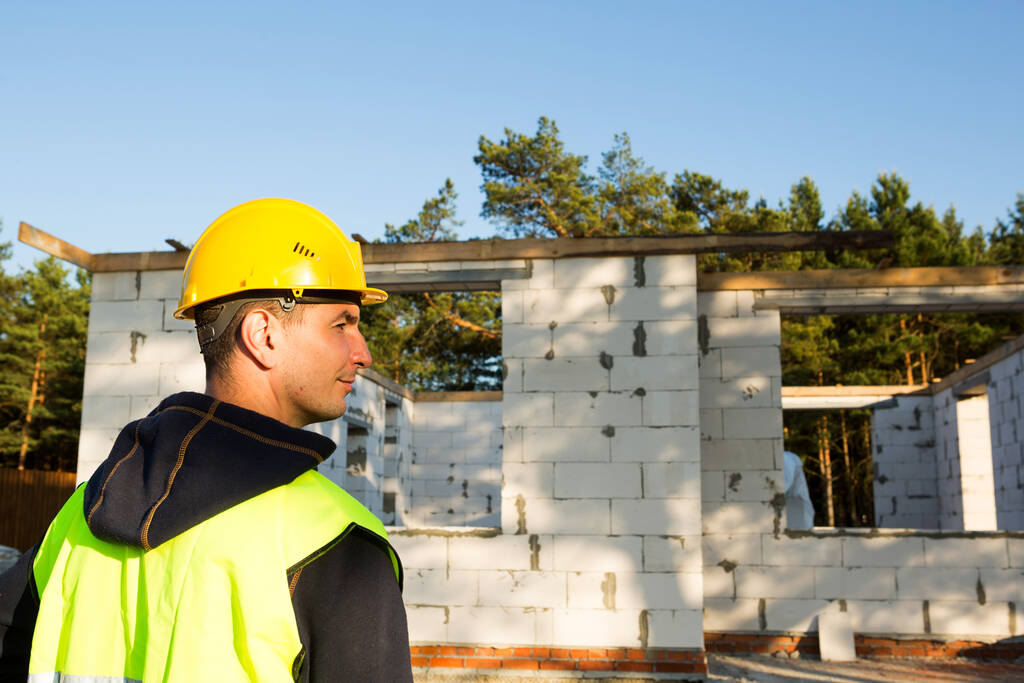 建築家は、壁を持つ家の建設現場の近くに立つ多孔質コンクリートブロックとレンガで作られています。保護黄色の硬い帽子と反射ベストの建設労働者. - 写真・画像