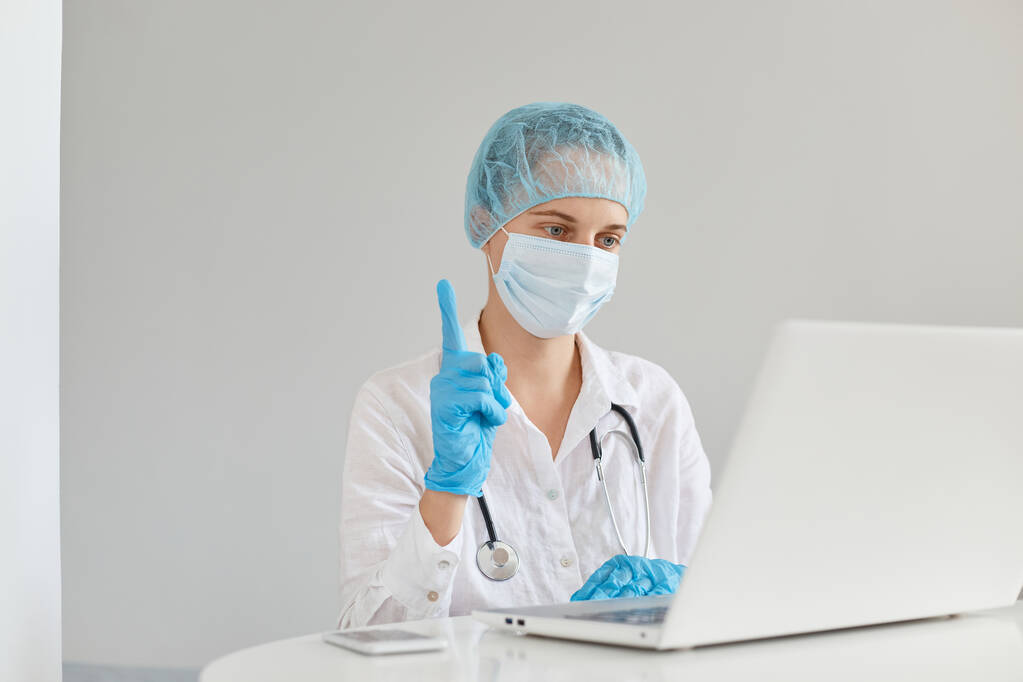 Női orvos terapeuta visel köpeny, sebészeti maszk, orvosi sapka és kesztyű, dolgozó számítógép előtt a klinikán, amelynek ötlete írni tudományos orvosi cikket, ujjal felfelé. - Fotó, kép