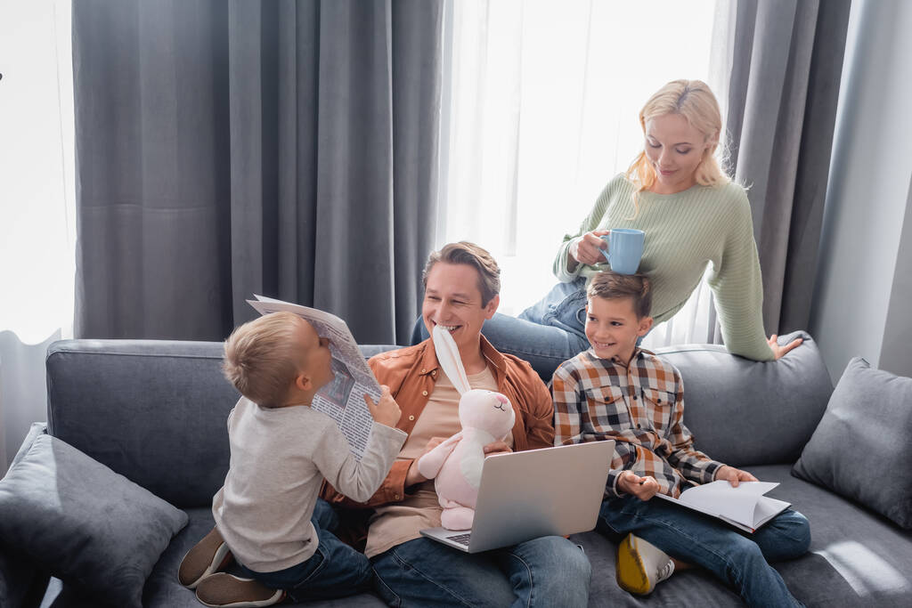 férfi laptoppal és játék nyuszi mosolygós közelében gyerekek és feleség csésze tea a nappaliban - Fotó, kép